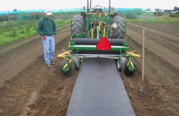 A machine lays plastic down a row of crop while a farmer observes.
