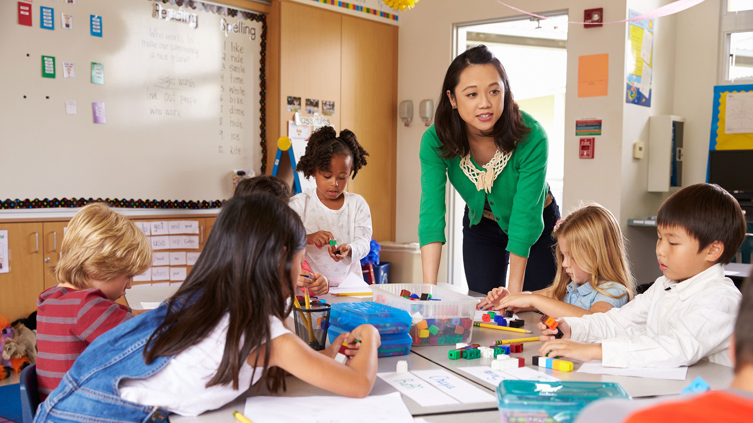 Elementary school students working at a table, with their teacher leaning over helping.