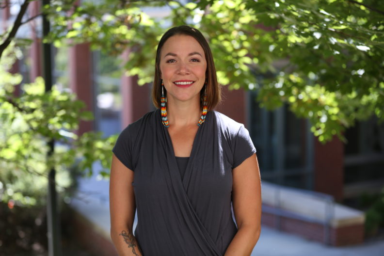 Stephany RunningHawk Johnson smiling at camera while standing in front of some trees blocking the way to the Education Addition building.