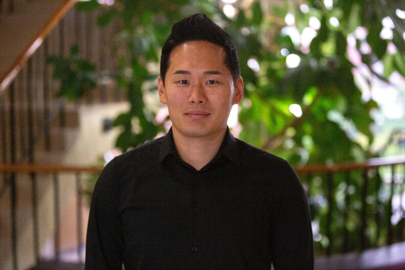 Alex Gang smiling in front of the ficus elastica in classy Cleveland Hall on a dark and dreary day on the Palouse, a byproduct of Hurricane Hilary coming up the coast line and pushing its way to the Inland Northwest.