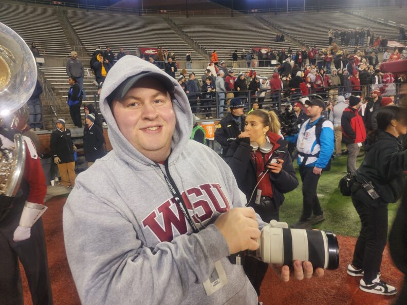 WSU ROAR scholar holding camera on sidelines at football game.