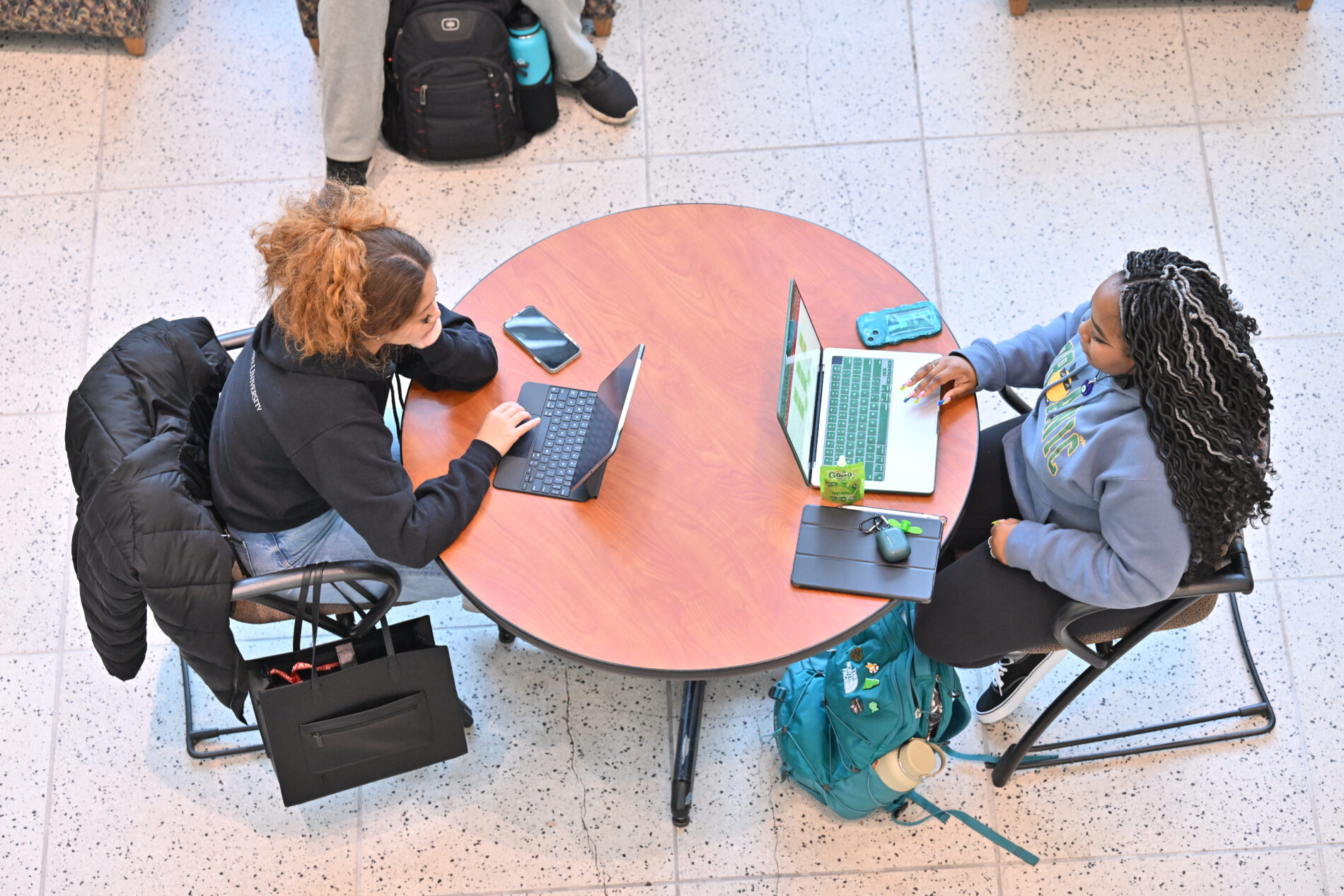 Two students sitting at a round table working on laptops in a campus study area.