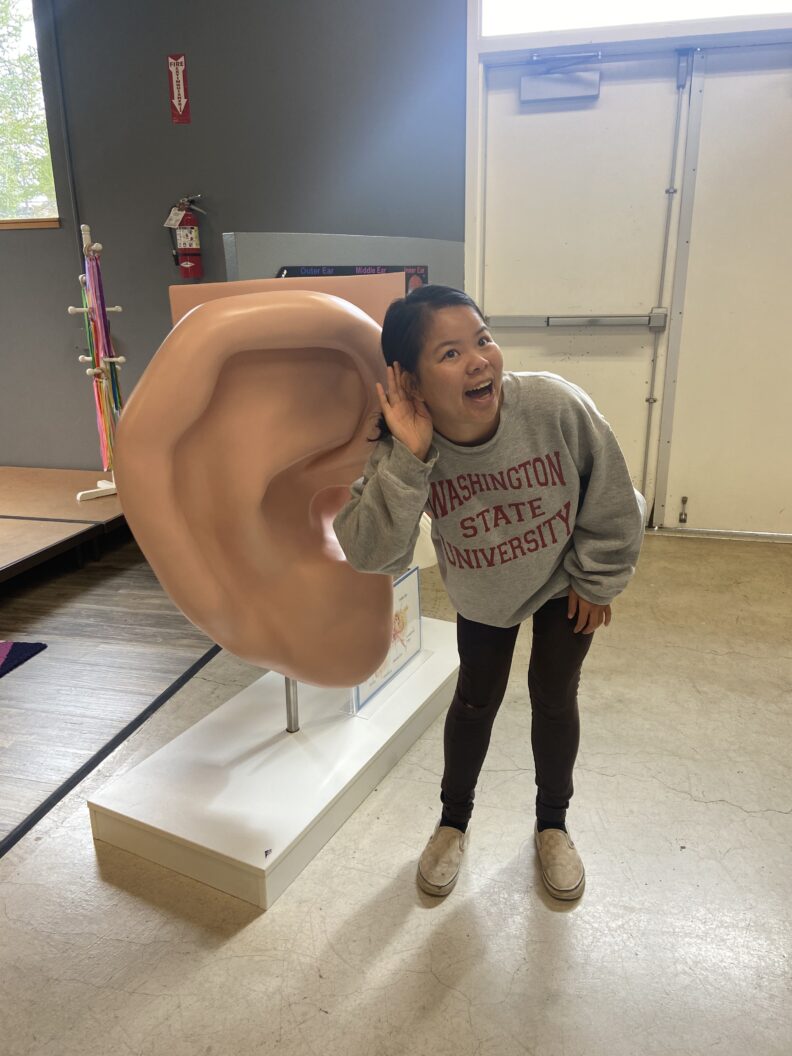 ROAR Scholar Emily Williamson holding her hand near her ear next to a very large ear sculpture at the Palouse Discover Science Center.