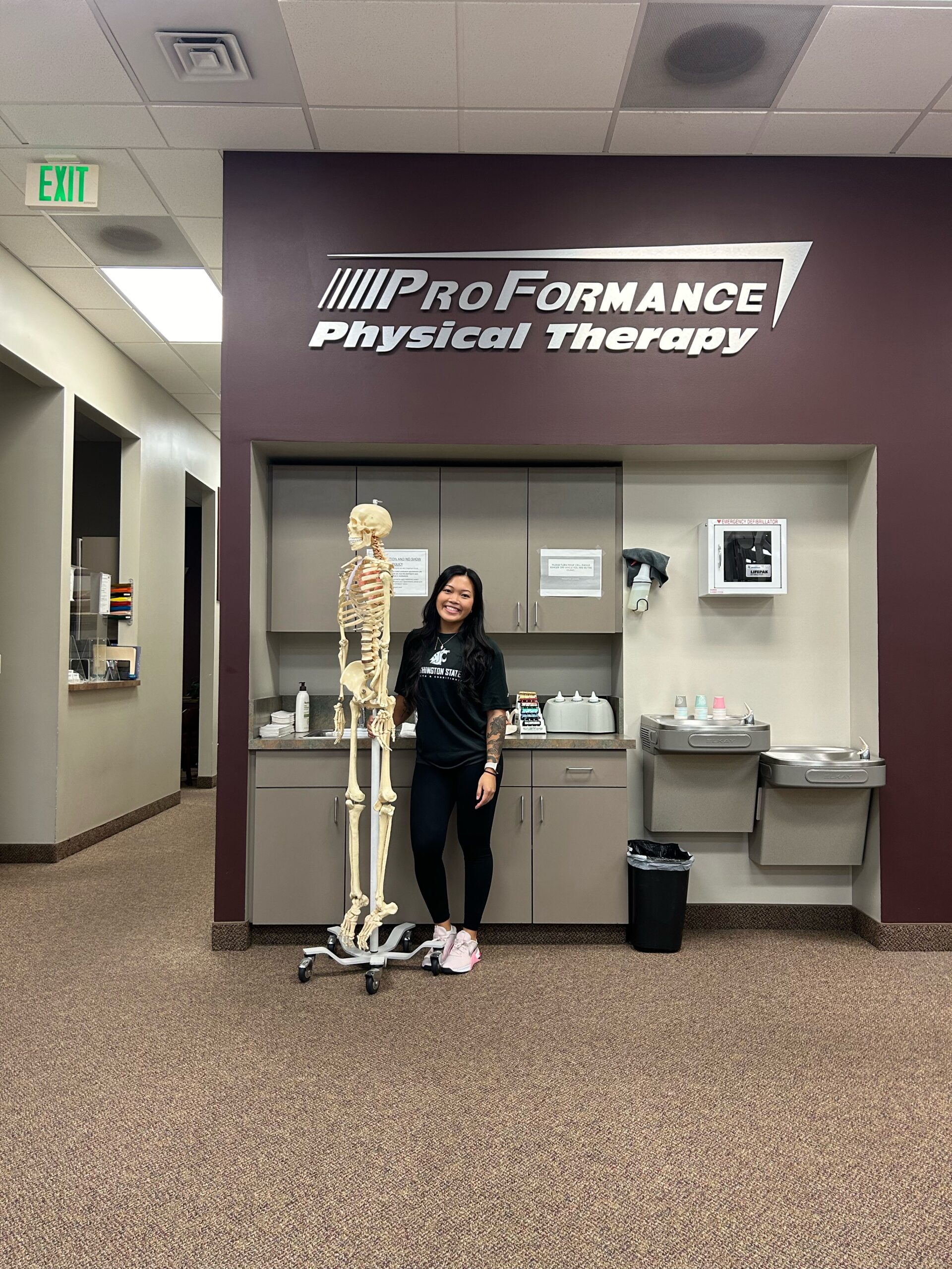 Individual standing beside a full-size human skeletal model in a physical therapy clinic setting
