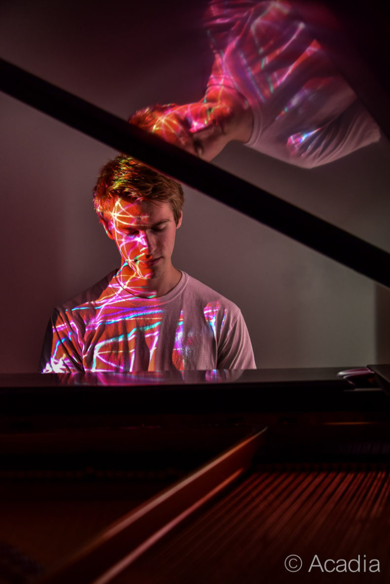A photo of a man playing a piano with his reflection on the lid of the piano