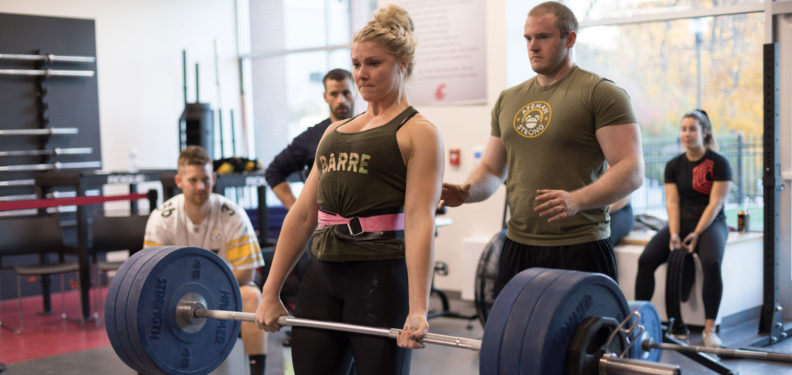 A girl lifting weights in a weight room while being spotted with a crowd of people watching