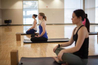 4 people in the room cross-legged on yoga mats with their eyes closed.