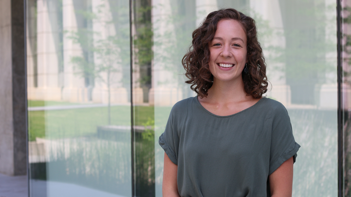 Tamika standing outside against a glass wall that is reflecting grass, trees, and benches.