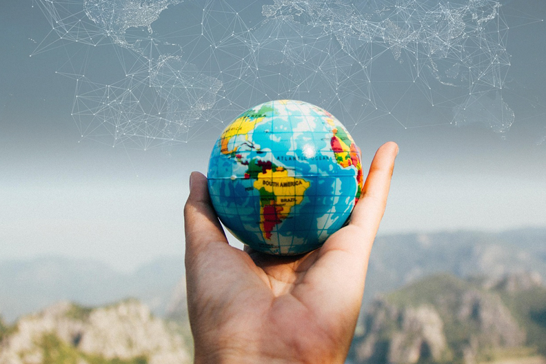 Hand holding model of the earth, with mountains in the background, and a map of the earth faintly in the sky.