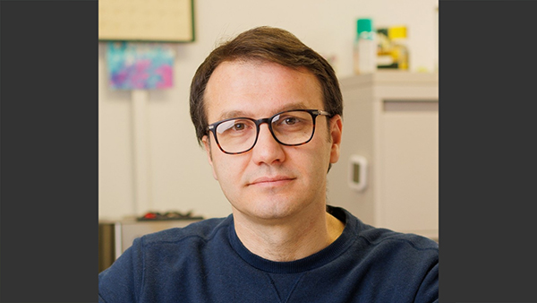Casual photo of Deniz in an office. Short brown hair and dark framed glasses.
