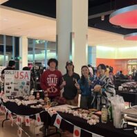 JSA members posing for a picture at a past event. They are standing at a decorated table at an event, which contains Japanese flags, games, and snacks.