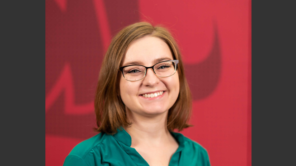 Chelsea, wearing a green blouse, and standing in front of a crimson wall with Coug head on it.