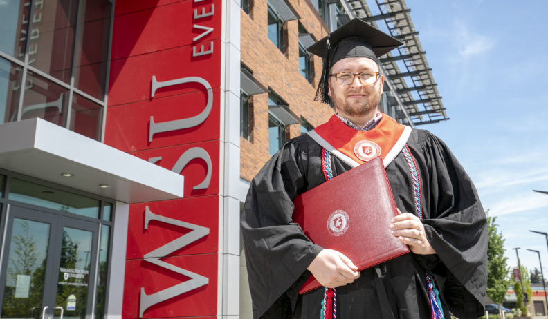 A graduating college student wearing graduation robes and holding a diploma standing in front of the WSU Everett building.