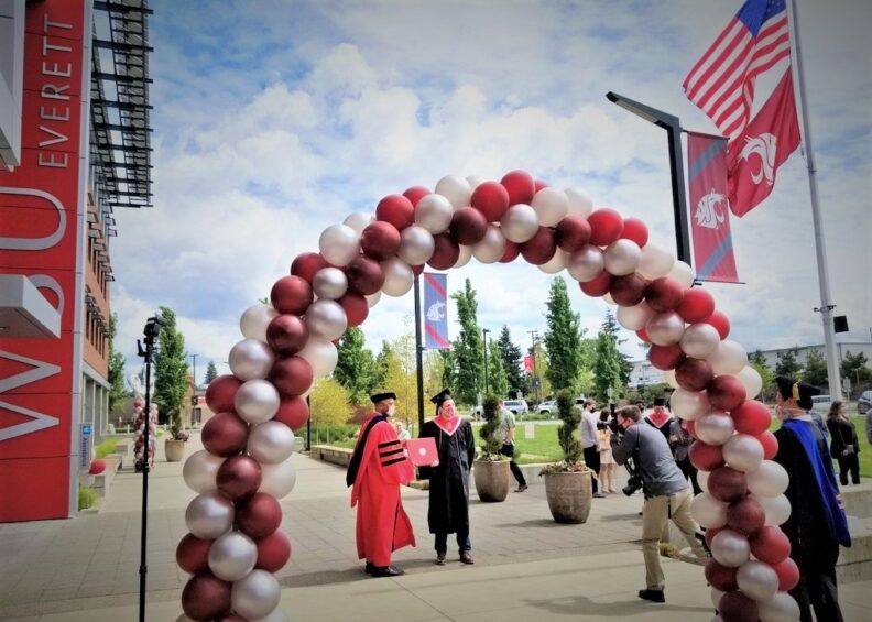 An archway made of crimson and white balloons with a college graduate underneath receiving a diploma.