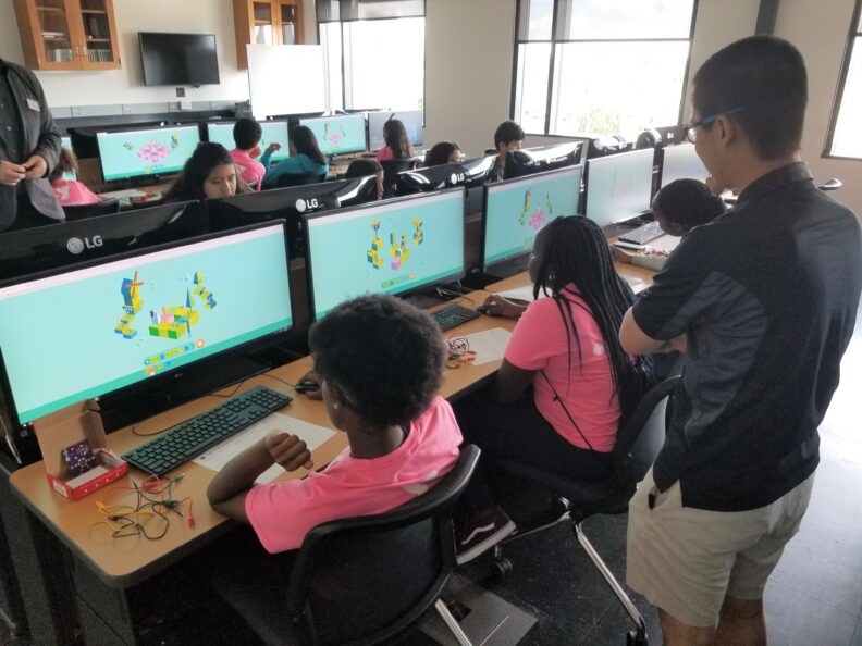 People working in a computer lab wearing matching pink shirts while others look on.