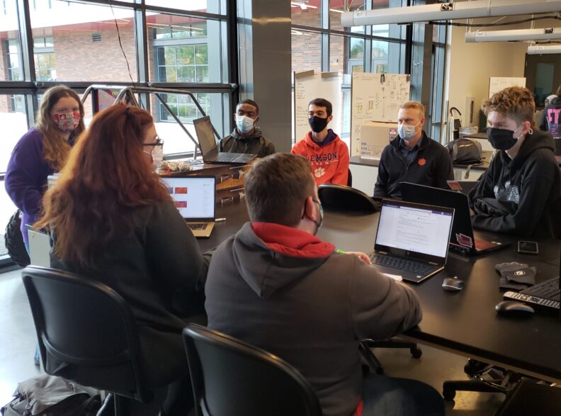 A group of people sitting around a table, many with laptops and wearing face masks.