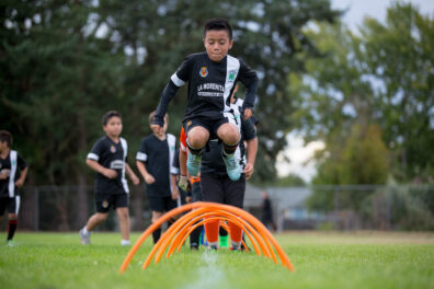 Children in soccer uniforms, jumping over hoops in a drill.