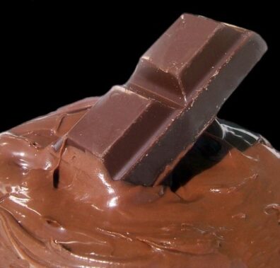 A block of chocolate sits in melted chocolate against a black backdrop.