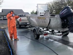 Man in orange protective suit sprays a metal boat on a trailer.