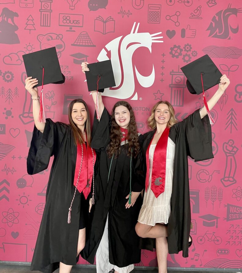 An image of three Brelsford Washington State University Visitor Center employees dressed in commencement regalia and cheering inside the facility.