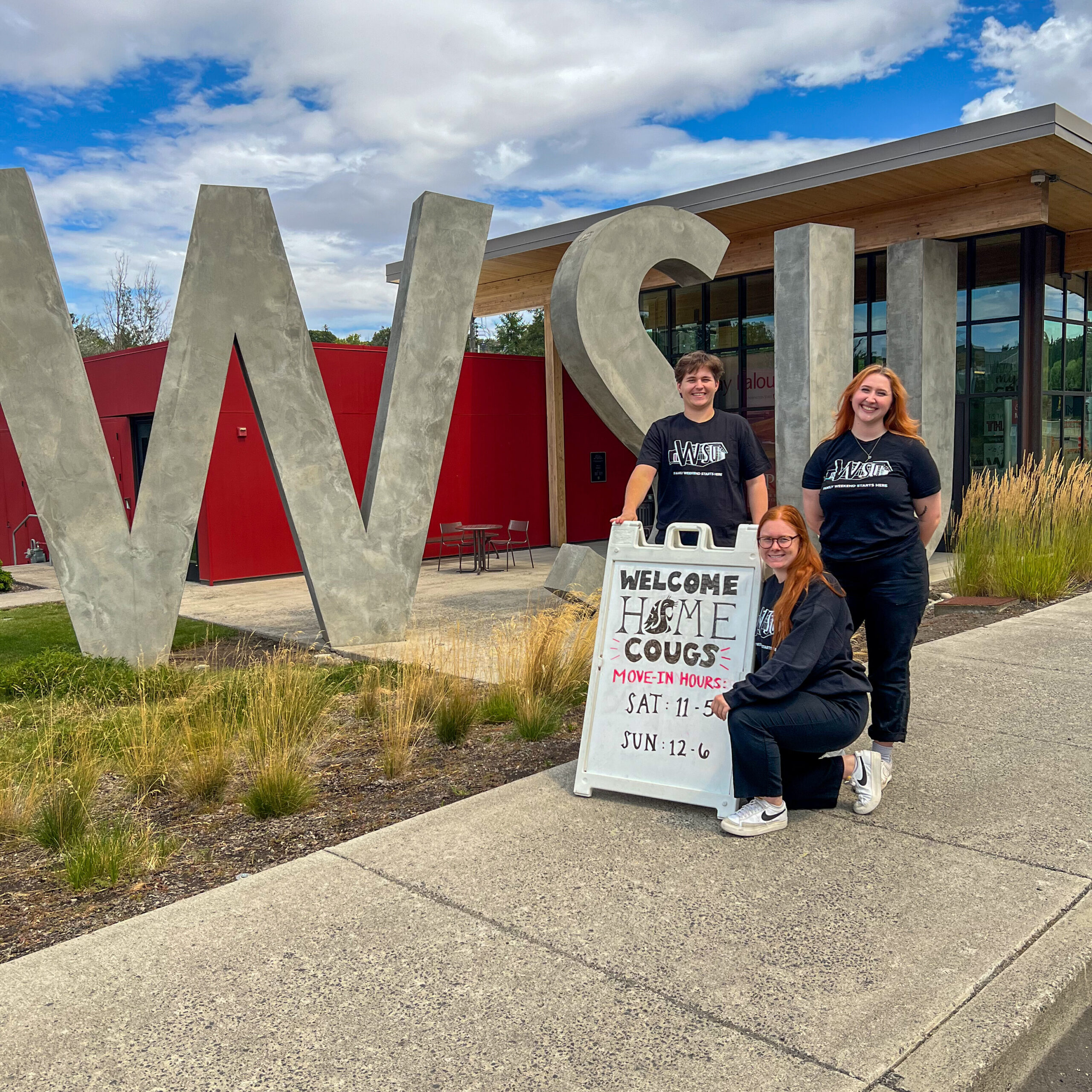 An image of a group of three Brelsford Washington State University Visitor Center employees around a sign for special Move-In Weekend hours.