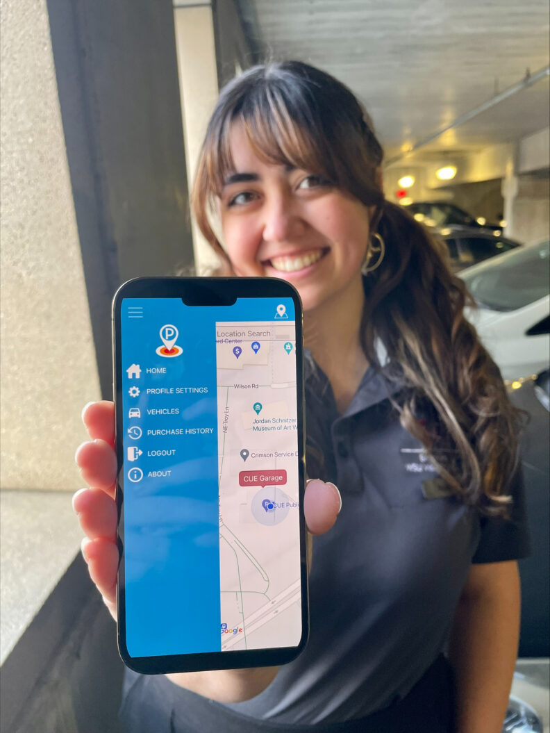 An image of a Brelsford Washington State University Visitor Center employee holding a phone displaying the AMP Park app inside a parking garage at WSU Pullman.
