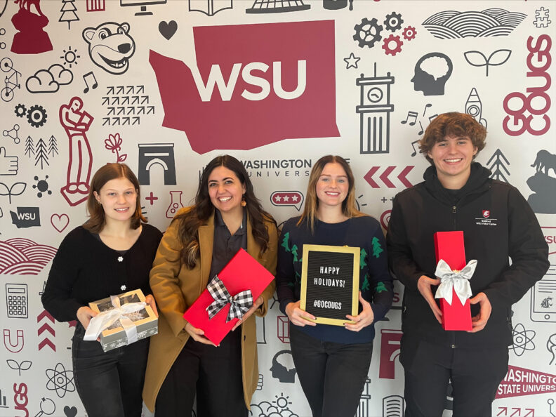 An image of four Brelsford Washington State University Visitor Center employees smiling together holding various Cougar-made gifts and a sign reading, "Happy Holidays!" inside the facility.