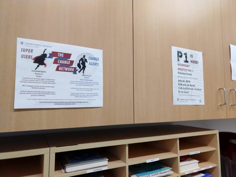 Two posters with white backgrounds stick to light brown wood cabinets above a matching set of wooden mailbox slots, some filled with pieces of mail. The left poster features a superhero next to the words Super User and a businessperson next to the words Change Agent, with smaller text below. The poster on the right reads P1 Prototype Preview with smaller text describing an upcoming event.