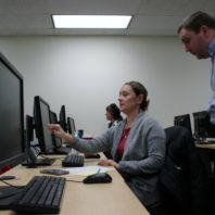We are looking down a long table at a row of computer monitors. A woman in a grey sweater points to her screen and talks to a man in a blue shirt who is looking over her shoulder at the computer.