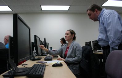 We are looking down a long table at a row of computer monitors. A woman in a grey sweater points to her screen and talks to a man in a blue shirt who is looking over her shoulder at the computer.