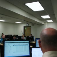 In the foreground, we see the back of a man's head as he stares at three computer screens. Past his monitors are rows of people at computers in a lab.