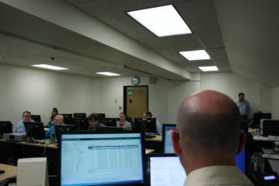In the foreground, we see the back of a man's head as he stares at three computer screens. Past his monitors are rows of people at computers in a lab.