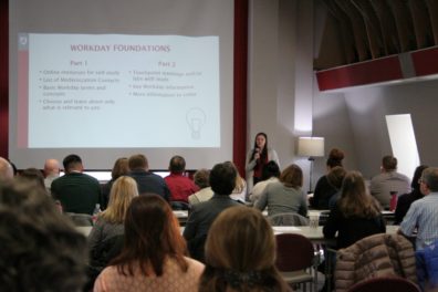 A presenter stands in front of the room with a group of Workday trainers listening.