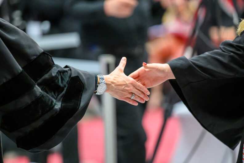 Close up photo of a handshake between two people wearing graduation gown. Image link leads to Executive Policy Manual page.