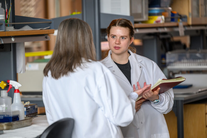 Photo of two people in lab coats, one holding a notebook, with shelves and cleaning supplies around them. Image link leads to Safety Policies and Procedures Manual page.