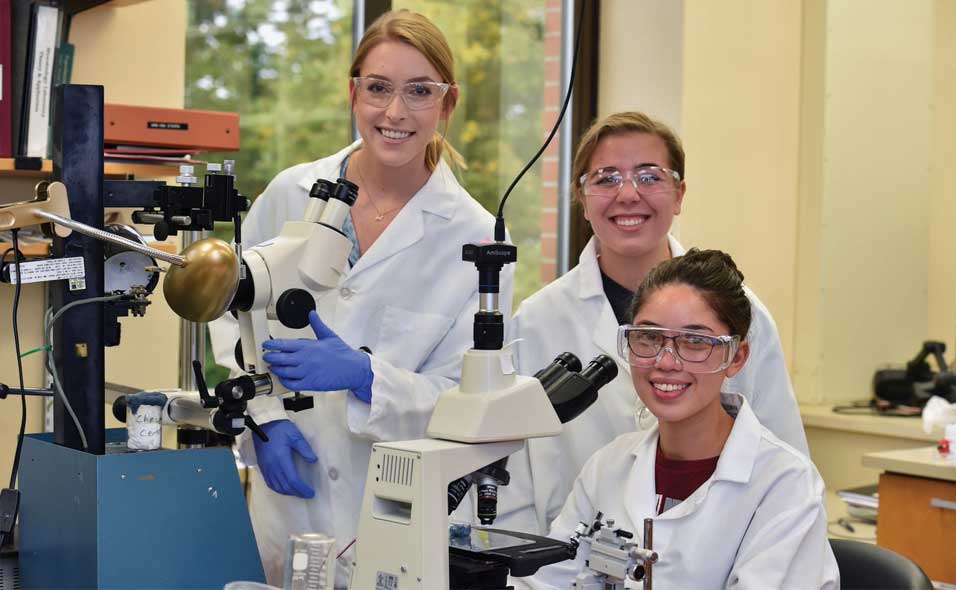Students in white lab coats examining samples under telescopes.