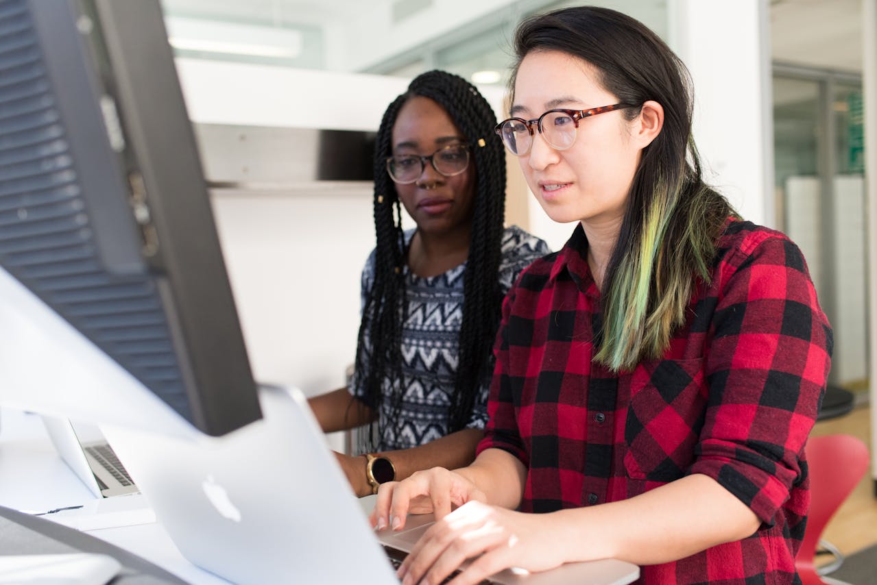 A student assists another student at a desktop computer.