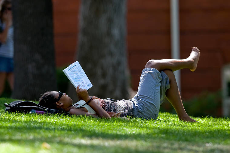A student lounges on the campus lawn and reads a book.