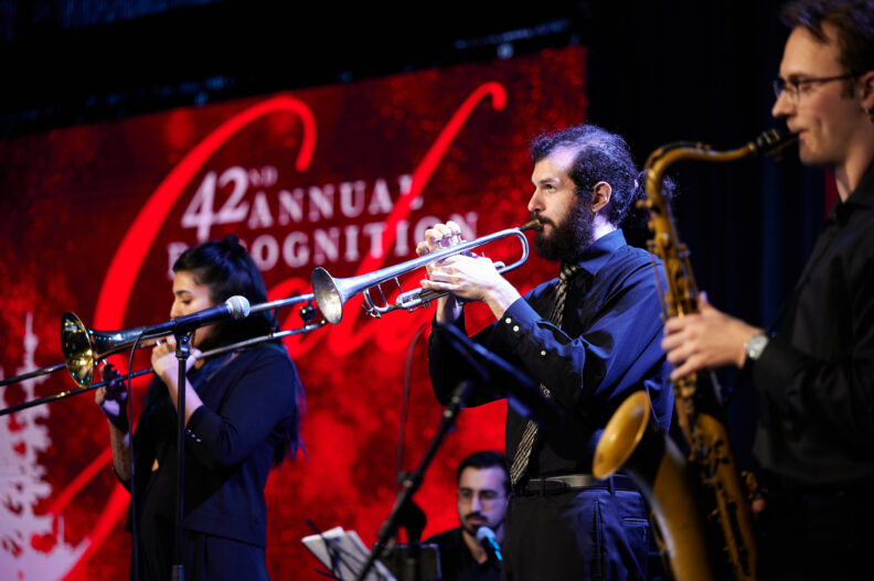 Band members play trumpets and a clarinet with the 42nd annual recognition gala logo screen in the background