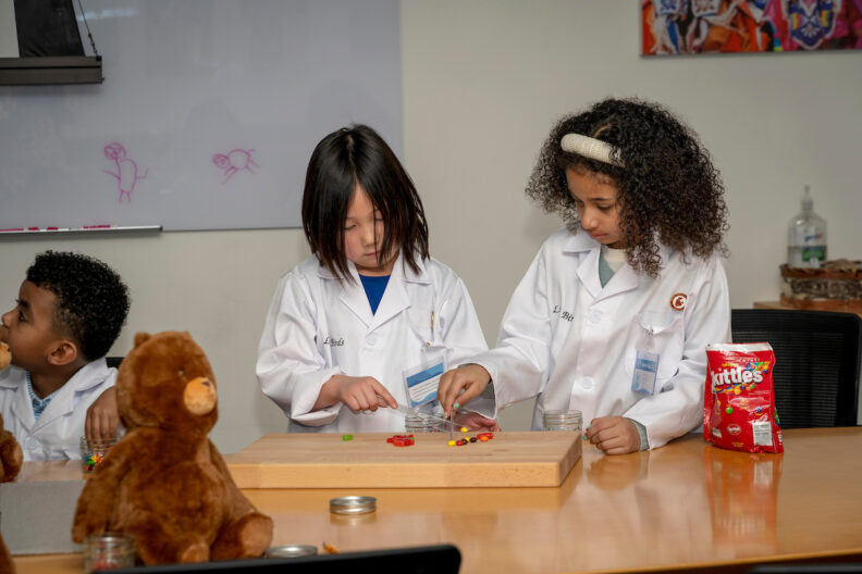 Two young children practice sorting skittles as stand-ins for pills while wearing Little Birds pharmacy jackets.