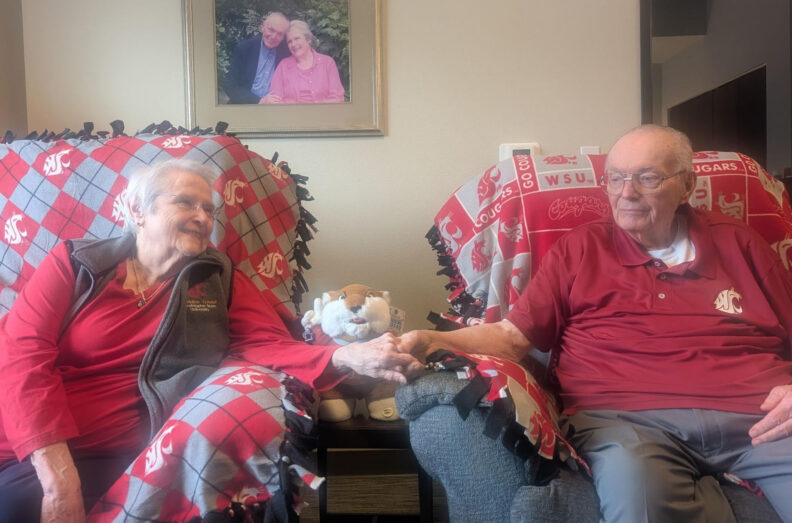 Gene and Priscilla Alberts sit side by side in their living room, holding hands while seated in armchairs draped with Washington State University blankets, with Cougar memorabilia visible between them.