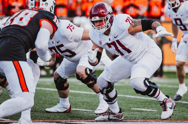 WSU football player Ashton Tripp (#51) in white and maroon uniform running with the ball during a game, helmet on, focused expression. Other players visible in background; stadium crowd in stands.