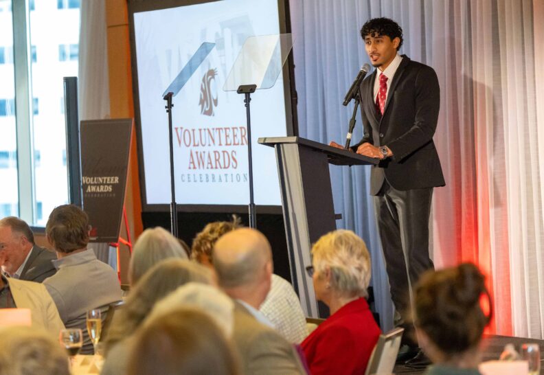 A student speaker in a suit and crimson tie addresses seated guests from a podium beneath the Volunteer Awards Celebration screen.