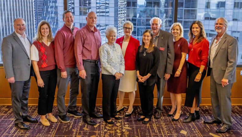 A group of eleven people, many dressed in crimson and gray, stand together for a photo in front of floor-to-ceiling windows overlooking a city skyline at the WSU Foundation's 2026 Volunteer Awards Celebration in Seattle.