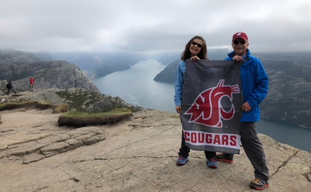 Claudia and John Swenson stand on a rocky mountain overlook holding a Washington State Cougars flag, with a dramatic fjord and steep cliffs stretching into the distance beneath cloudy skies.