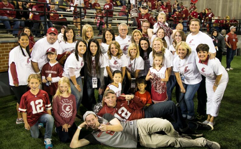 A large group of Washington State University alumni, family members, and children pose together on the field during a WSU football game, wearing Cougar apparel and alumni shirts, with fans filling the stadium stands in the background.