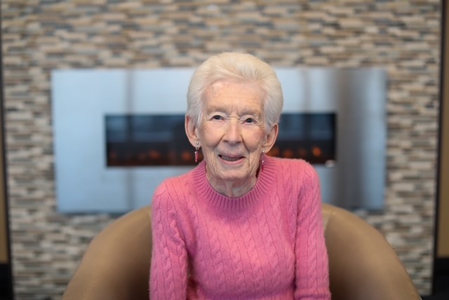 Portrait of a person seated in a chair wearing a pink sweater, with a contemporary fireplace and stone-tile wall in the background.
