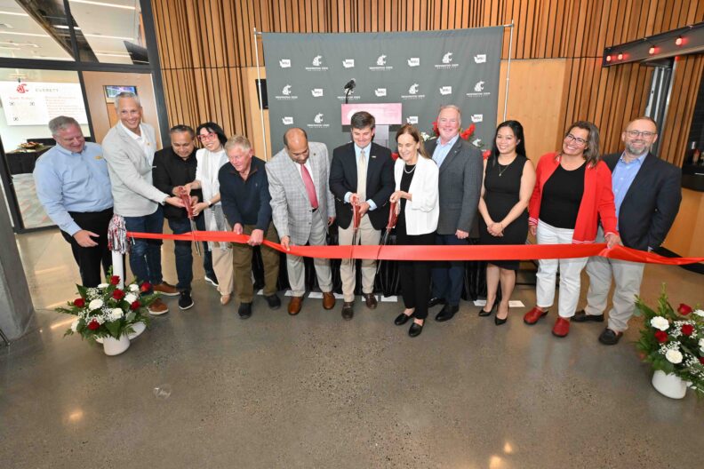 A line of people hold a red ribbon while two people with large scissors cut the ribbon at the Fluke Lab ribbon cutting.