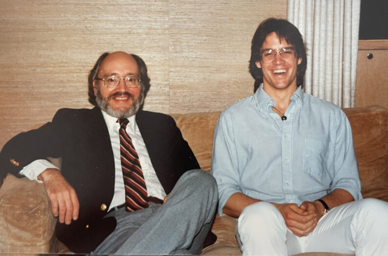 Dr. Roy Filby and another person sit together on a couch in a living room.
