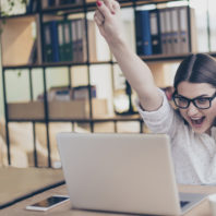Photo: Young woman sitting at laptop raises her clenched fists over her head in triumph.